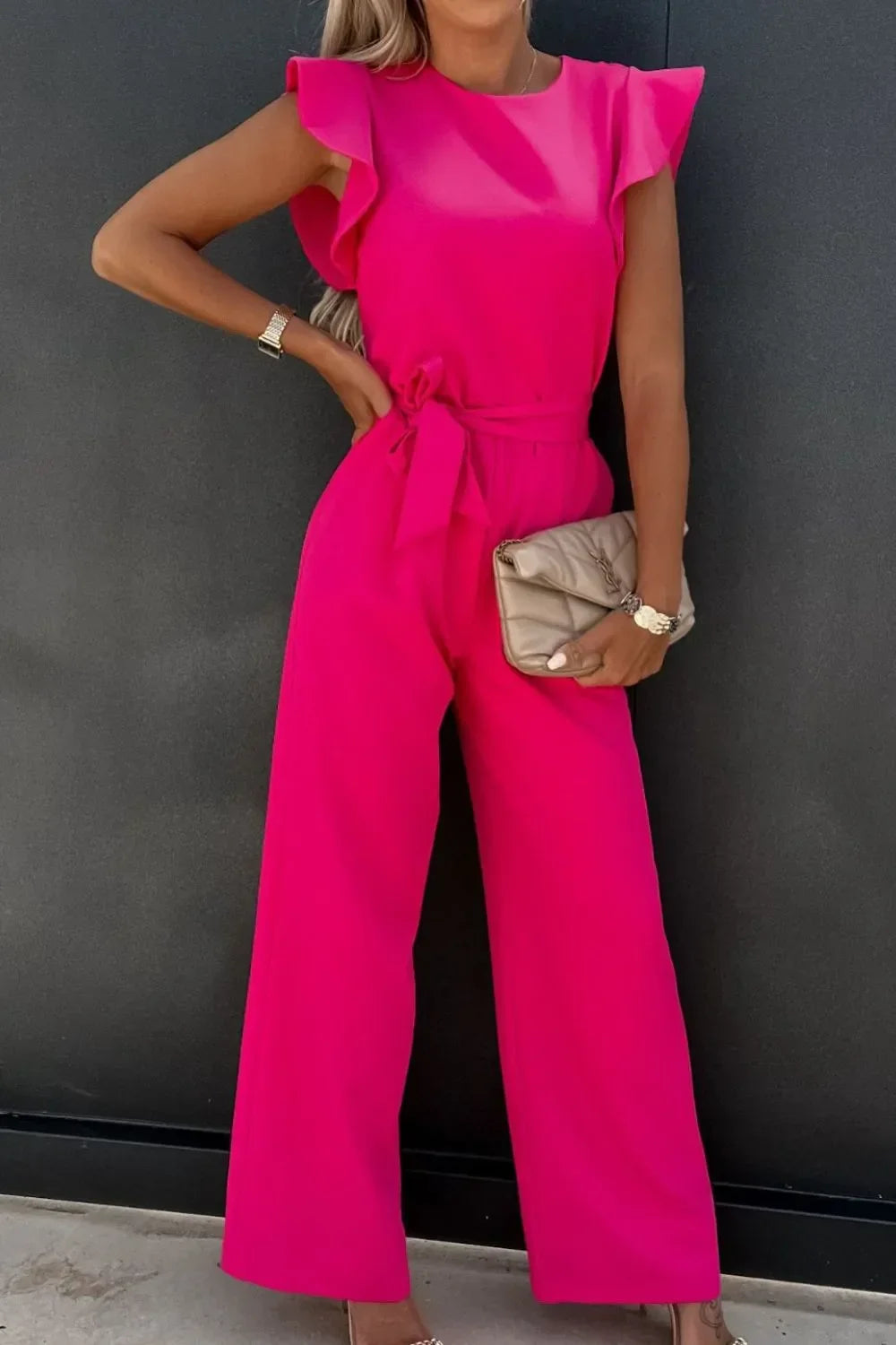 Woman wearing bright pink wide-leg jumpsuit with ruffled sleeves and waist tie, holding beige quilted clutch