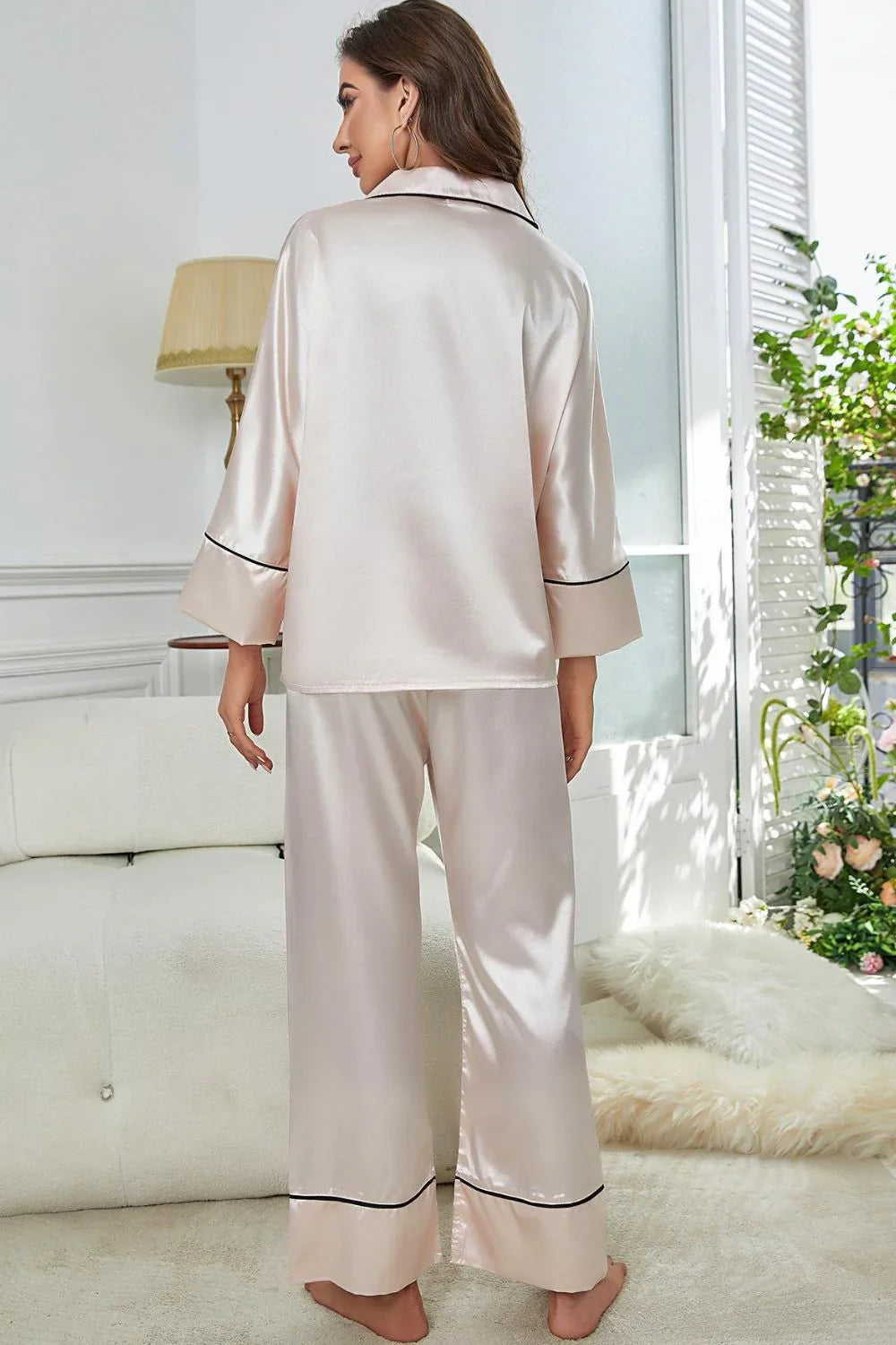 Woman in light pink satin pajama set with black piping standing barefoot in cozy room