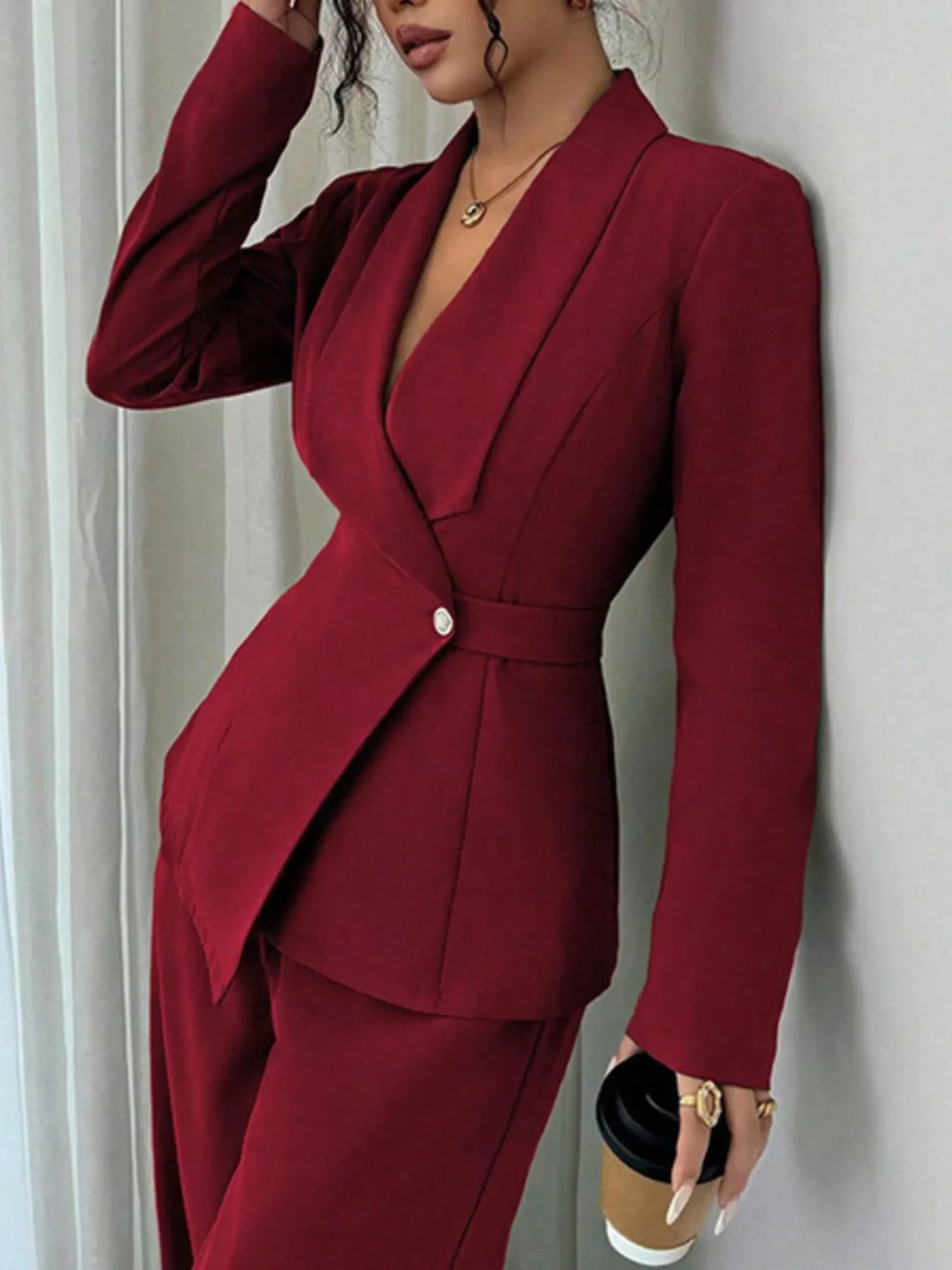 Woman in tailored burgundy pantsuit with V-neck blazer, holding a coffee cup indoors