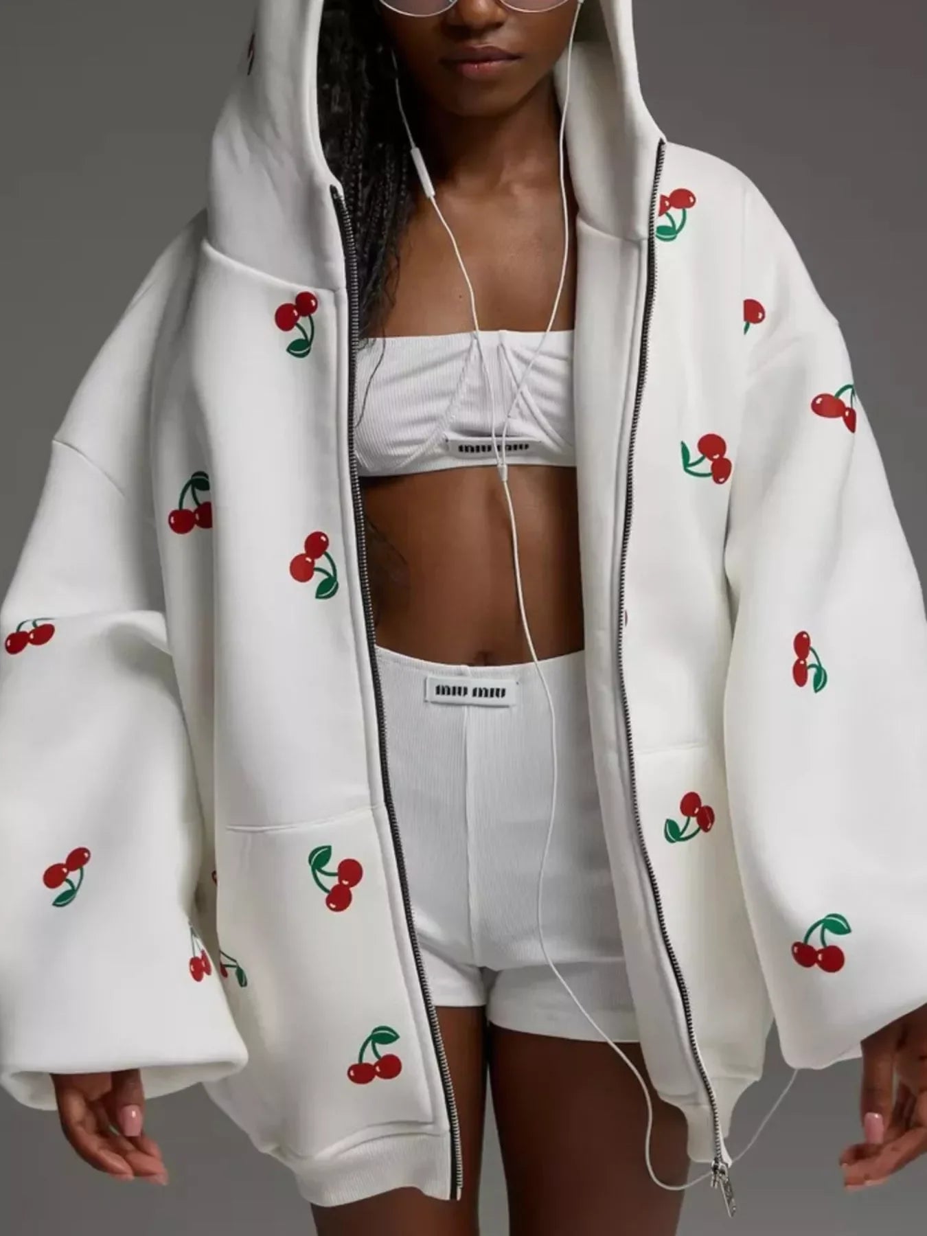 Woman wearing oversized white zip-up hoodie with red cherry prints and white sports bra and shorts