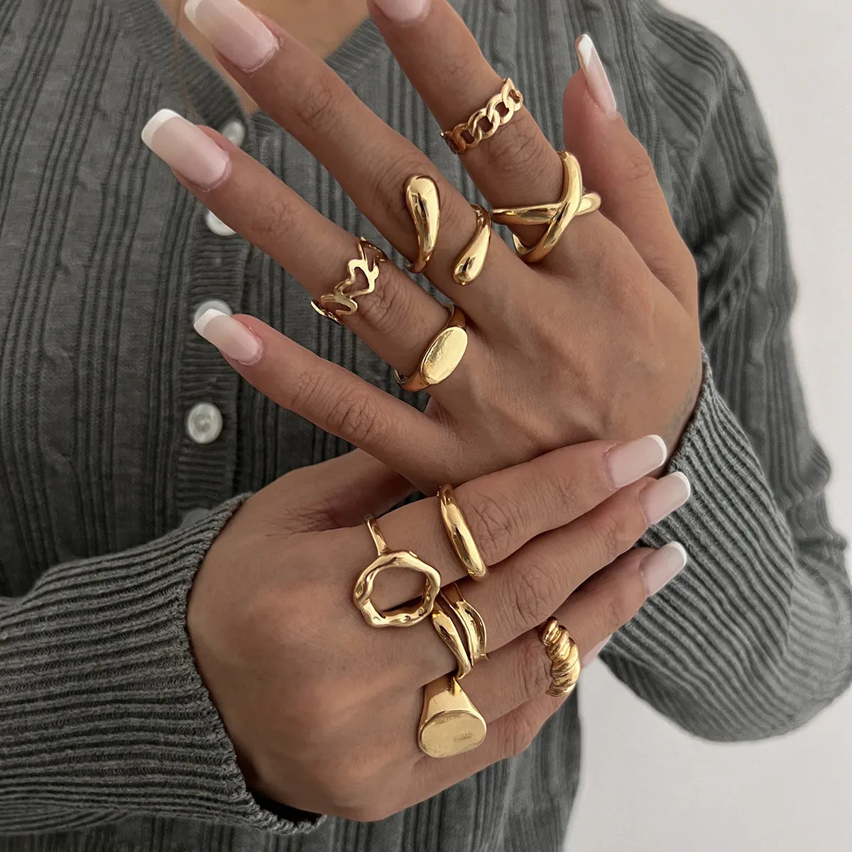 Gold statement rings set on hands with French manicure, styled with gray ribbed sweater for everyday elegance