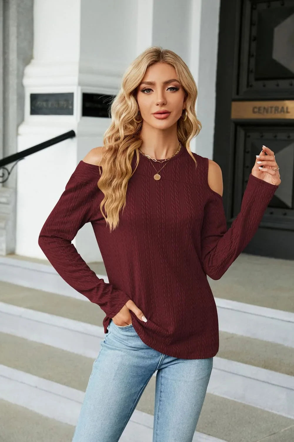 Woman in a burgundy cold shoulder sweater and light wash jeans standing on outdoor steps