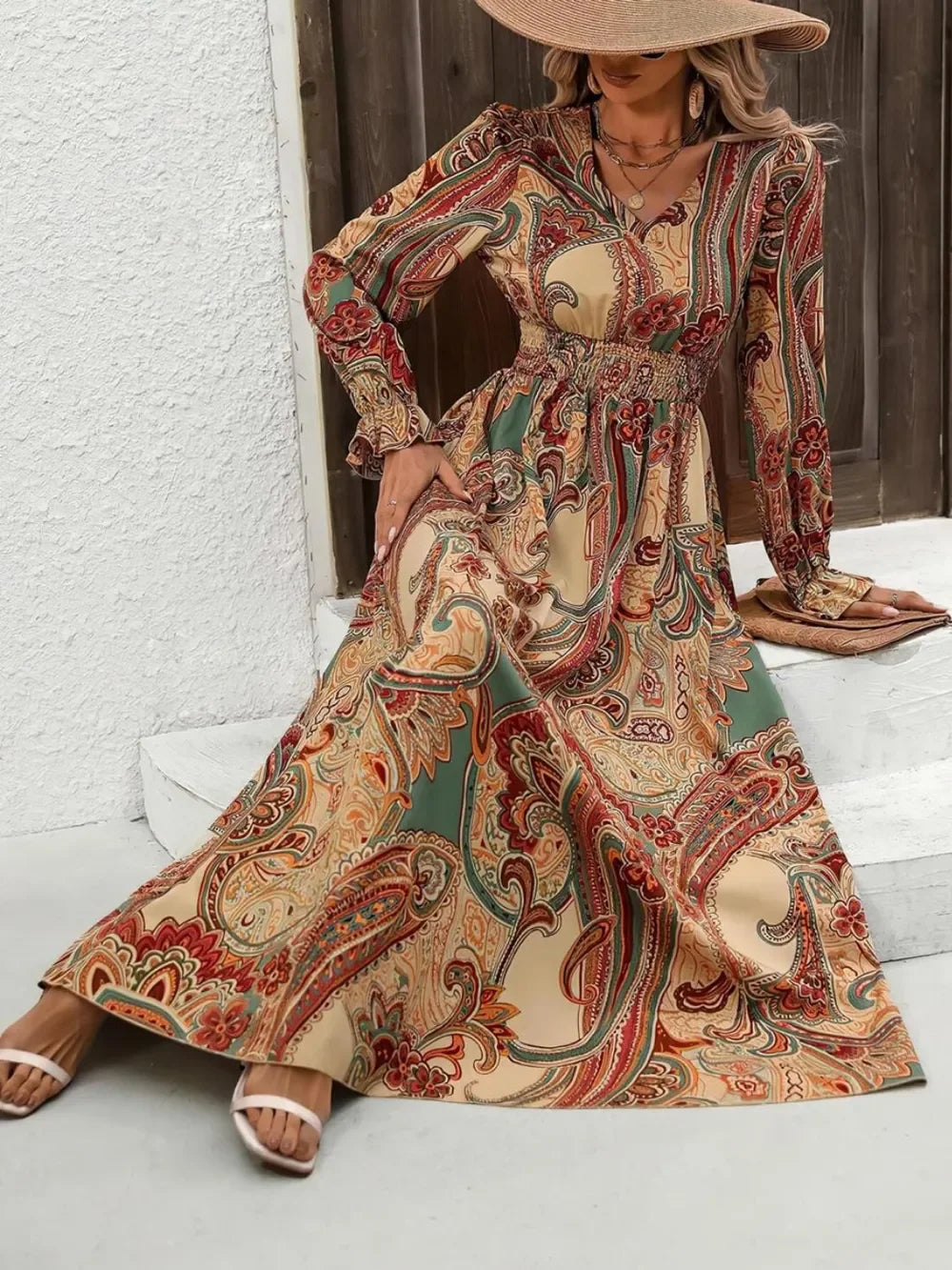 Woman in long paisley print boho maxi dress with smocked waist, wide-brim hat, and white sandals sitting on steps
