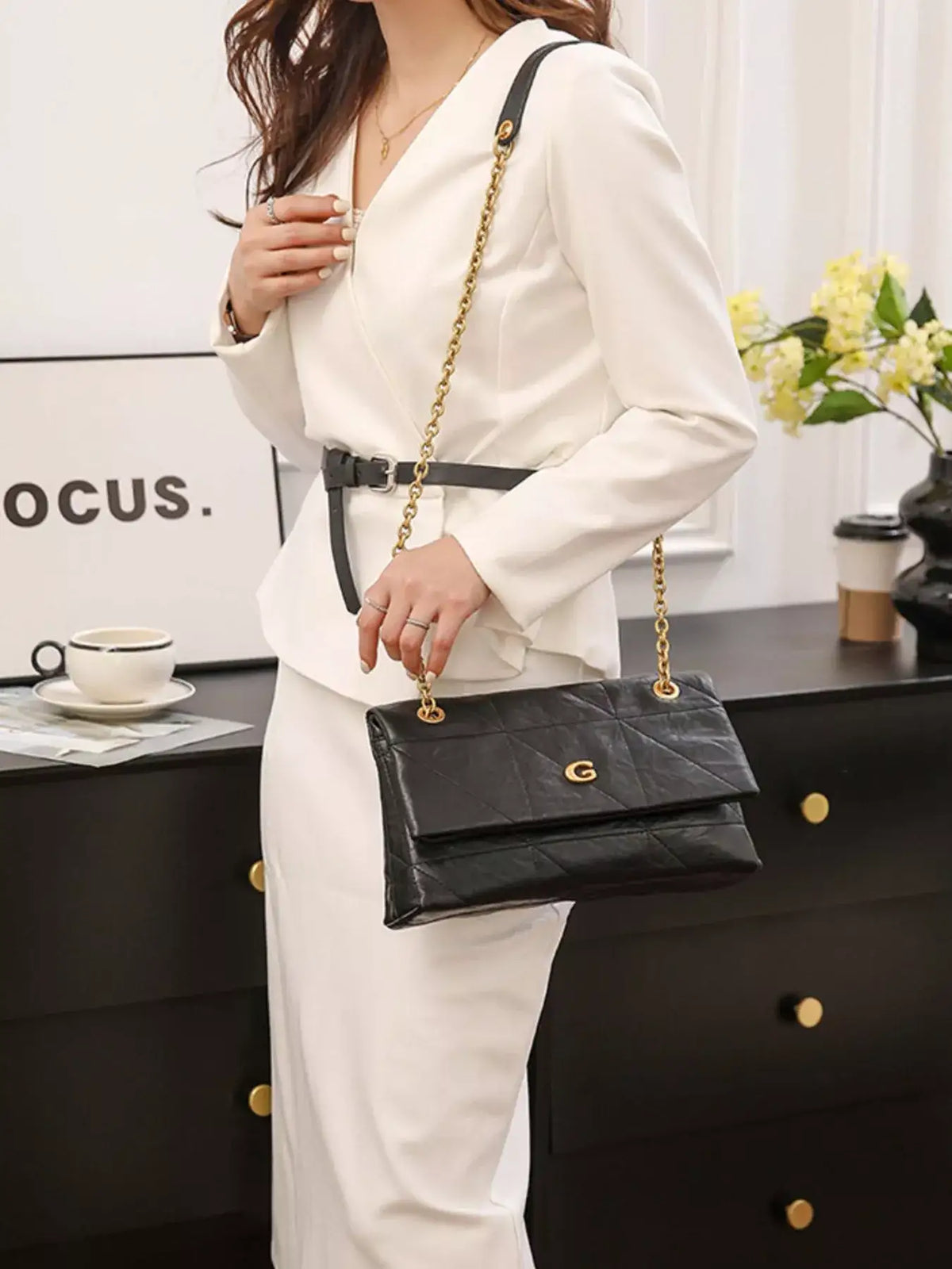 Woman in a white blazer set wearing a black geometric quilted shoulder bag with a gold chain strap.
