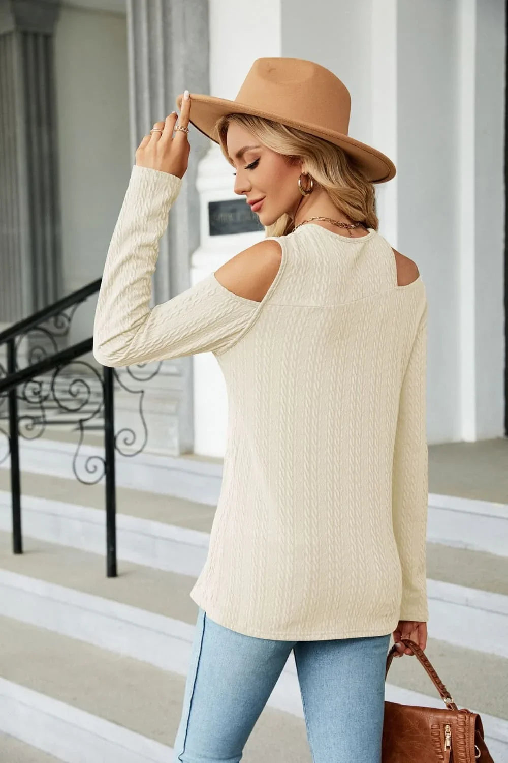 Woman in beige cold shoulder knit sweater, tan hat, and blue jeans holding a brown handbag.