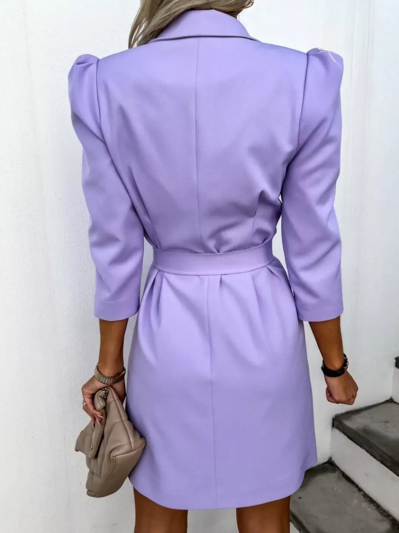 Back view of woman wearing lavender belted blazer dress with puffed shoulders and holding beige clutch