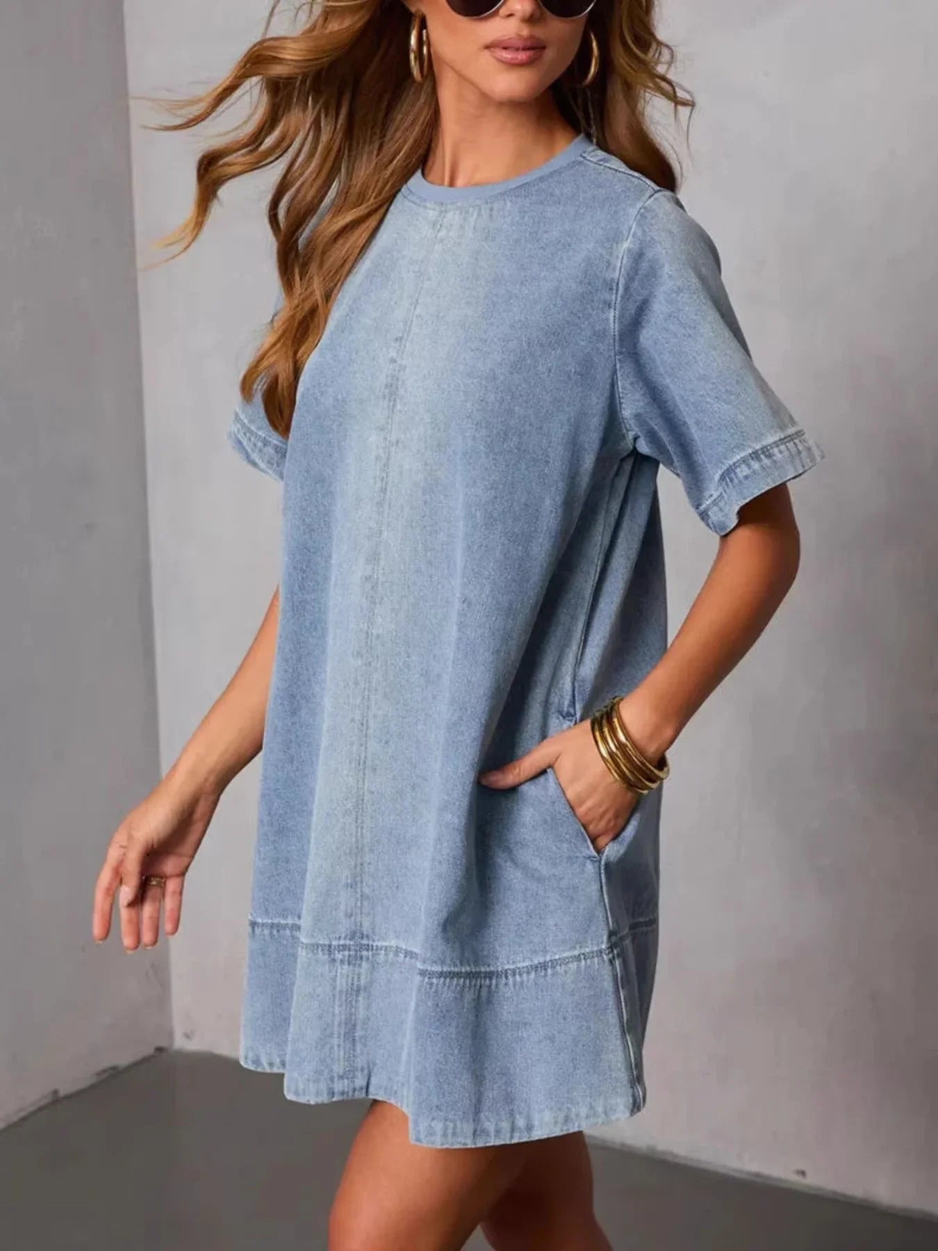 Woman wearing light blue denim short-sleeve dress with pockets, gold bangles, and sunglasses