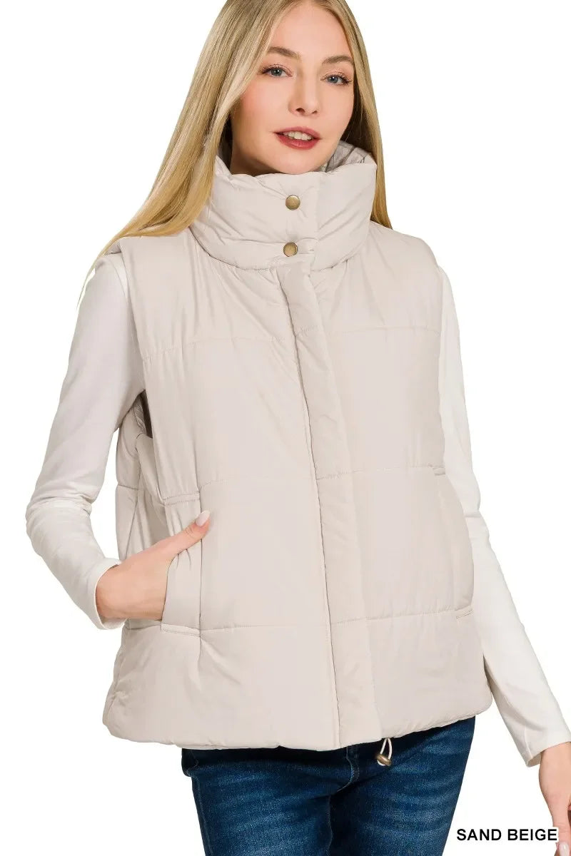 Woman wearing sand beige puffer vest with high collar and snap buttons, styled with long sleeve shirt and jeans