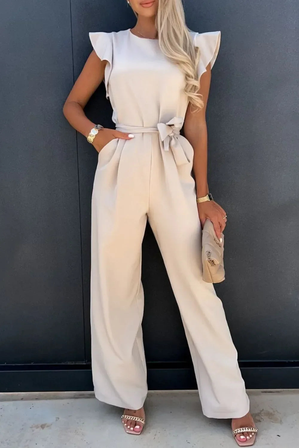 Woman wearing beige sleeveless wide-leg jumpsuit with ruffle details, carrying clutch against dark wall