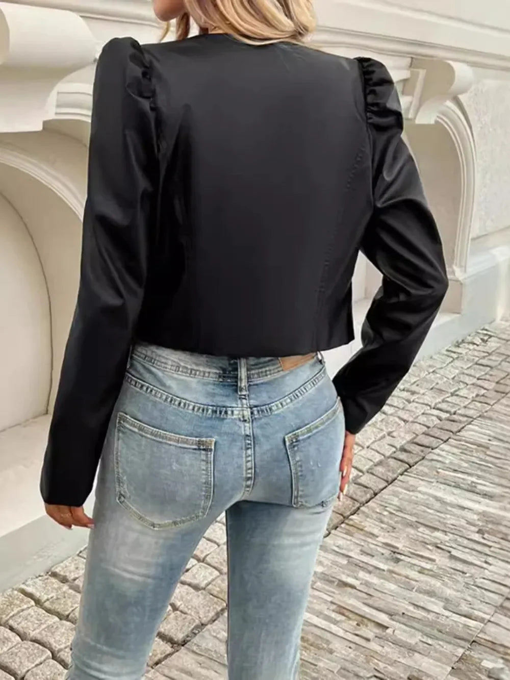 Back view of a woman wearing a black cropped jacket with puffed sleeves and light blue jeans.