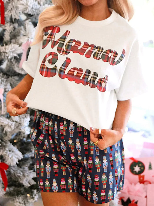 Woman wearing white Mama Claus T-shirt with plaid lettering and nutcracker-themed shorts by Christmas tree
