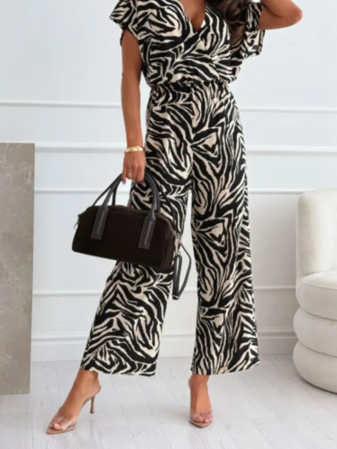 Woman wearing black and white zebra print jumpsuit holding black handbag and wearing clear heels