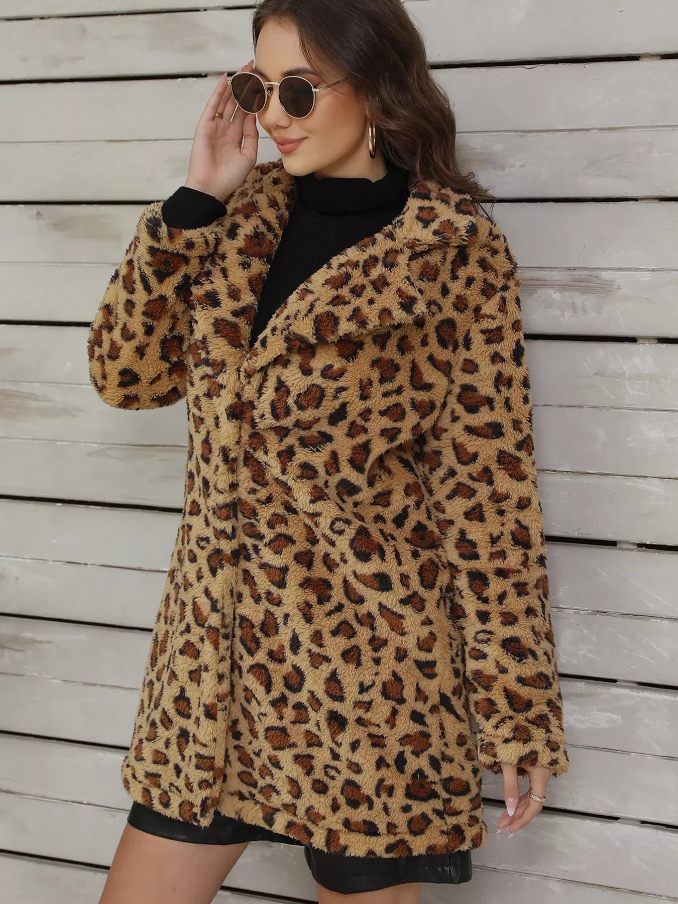 Woman wearing leopard print faux fur coat with round sunglasses and black turtleneck against wooden wall