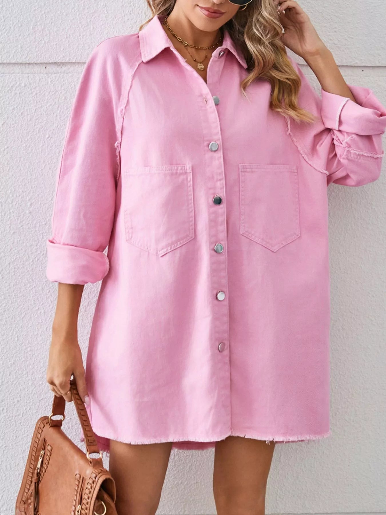 Woman wearing oversized pink button-down shirt dress with long sleeves and front pockets, holding tan handbag.