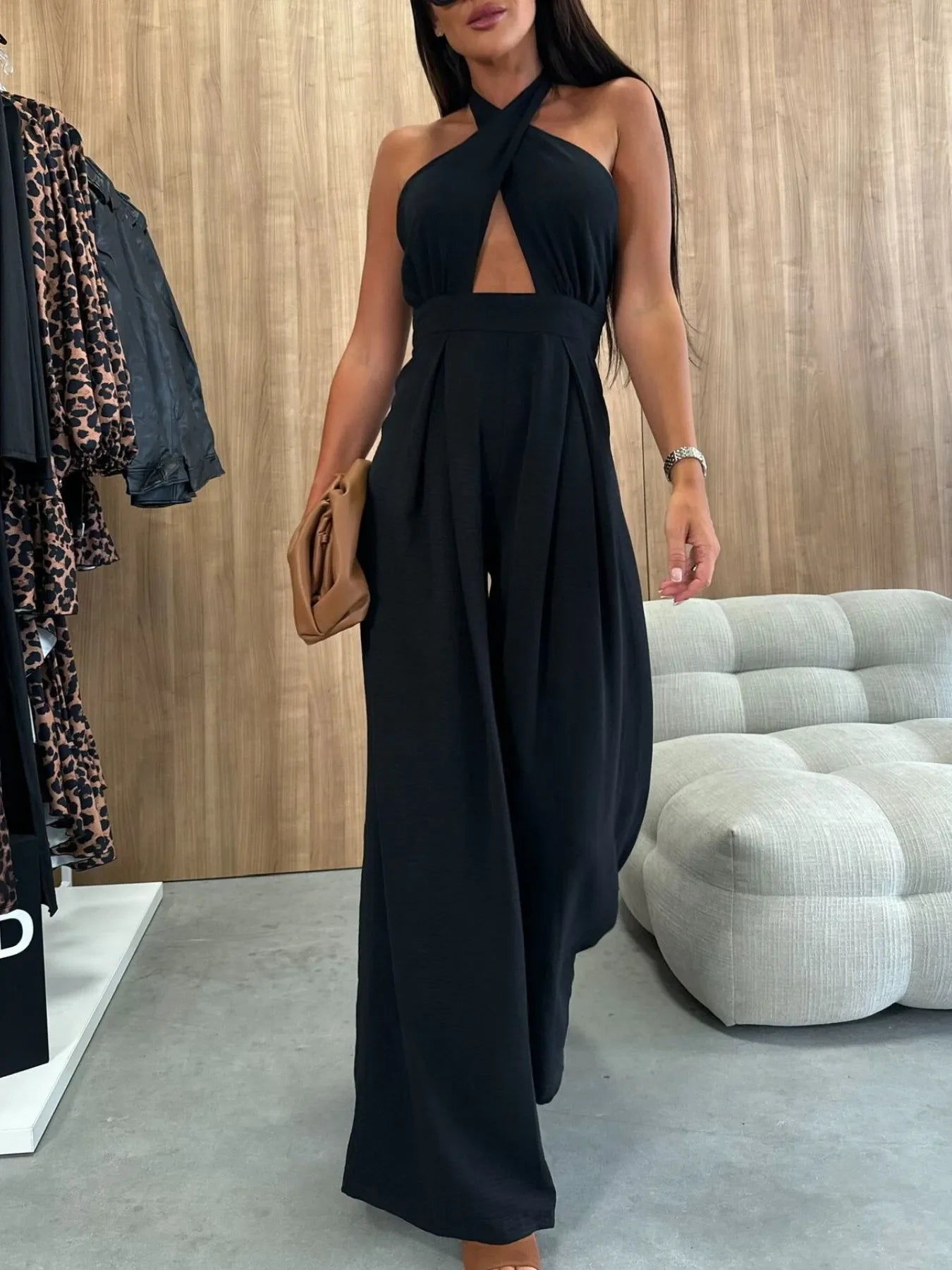Woman wearing black halter neck wide-leg jumpsuit holding brown clutch in modern indoor setting