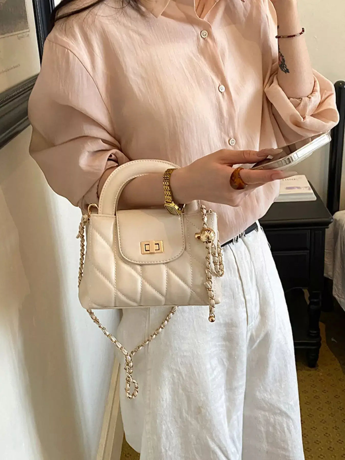 Woman wearing a beige blouse and white pants holding a quilted cream PU leather chain strap crossbody bag
