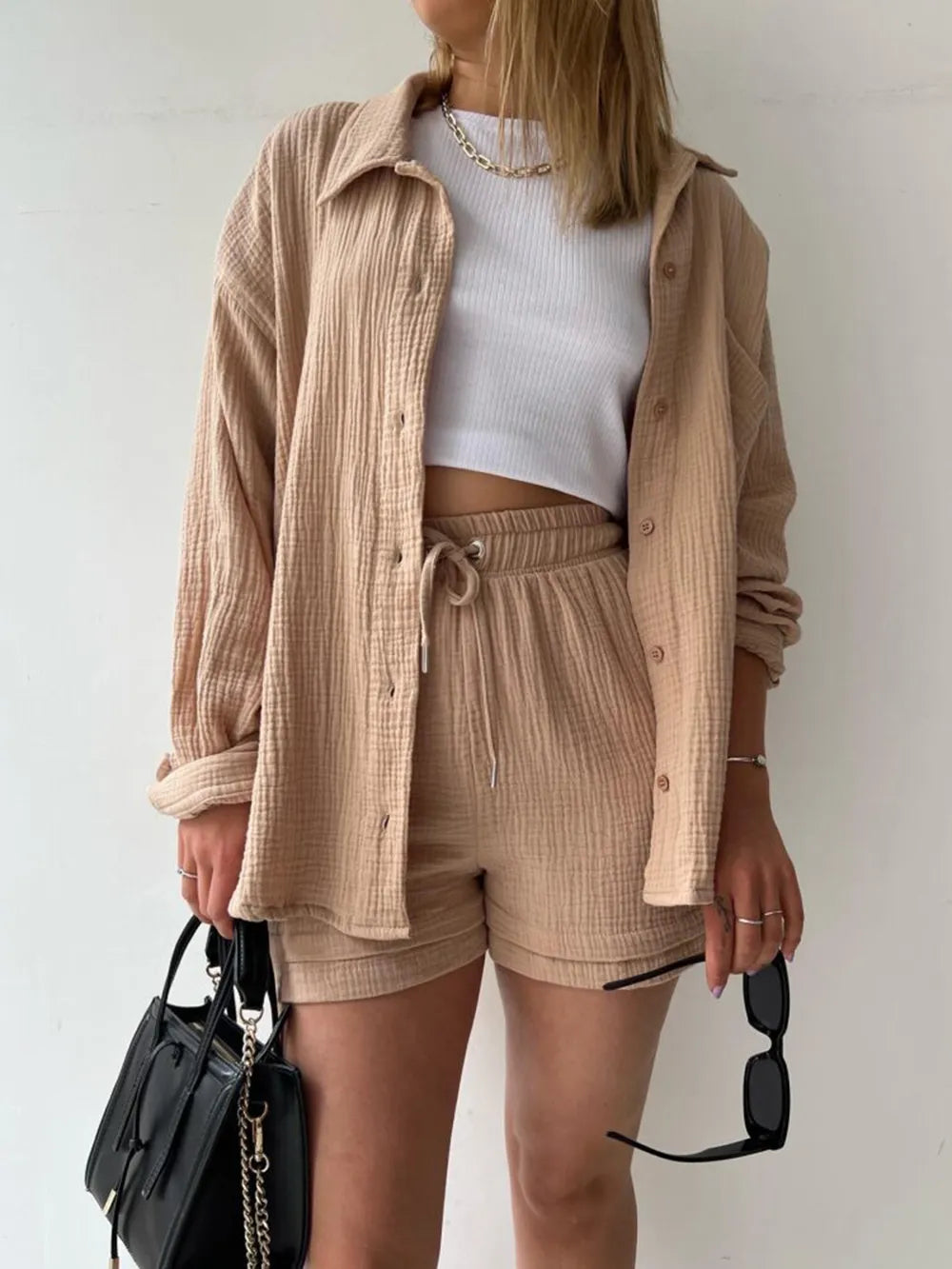 Woman wearing beige textured shorts set, white crop top, black handbag, holding sunglasses