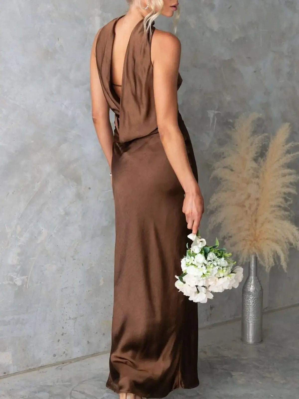 Back view of a brown satin maxi dress by Evie's Dreamland with a draped open back, paired with white flowers.