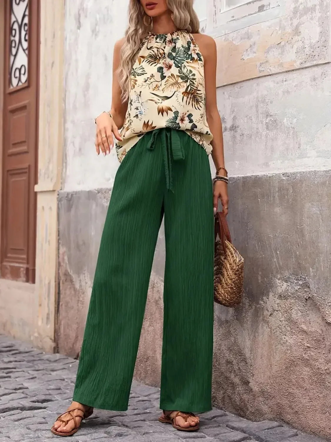 Woman wearing sleeveless floral print blouse and green wide-leg pants, holding straw bag