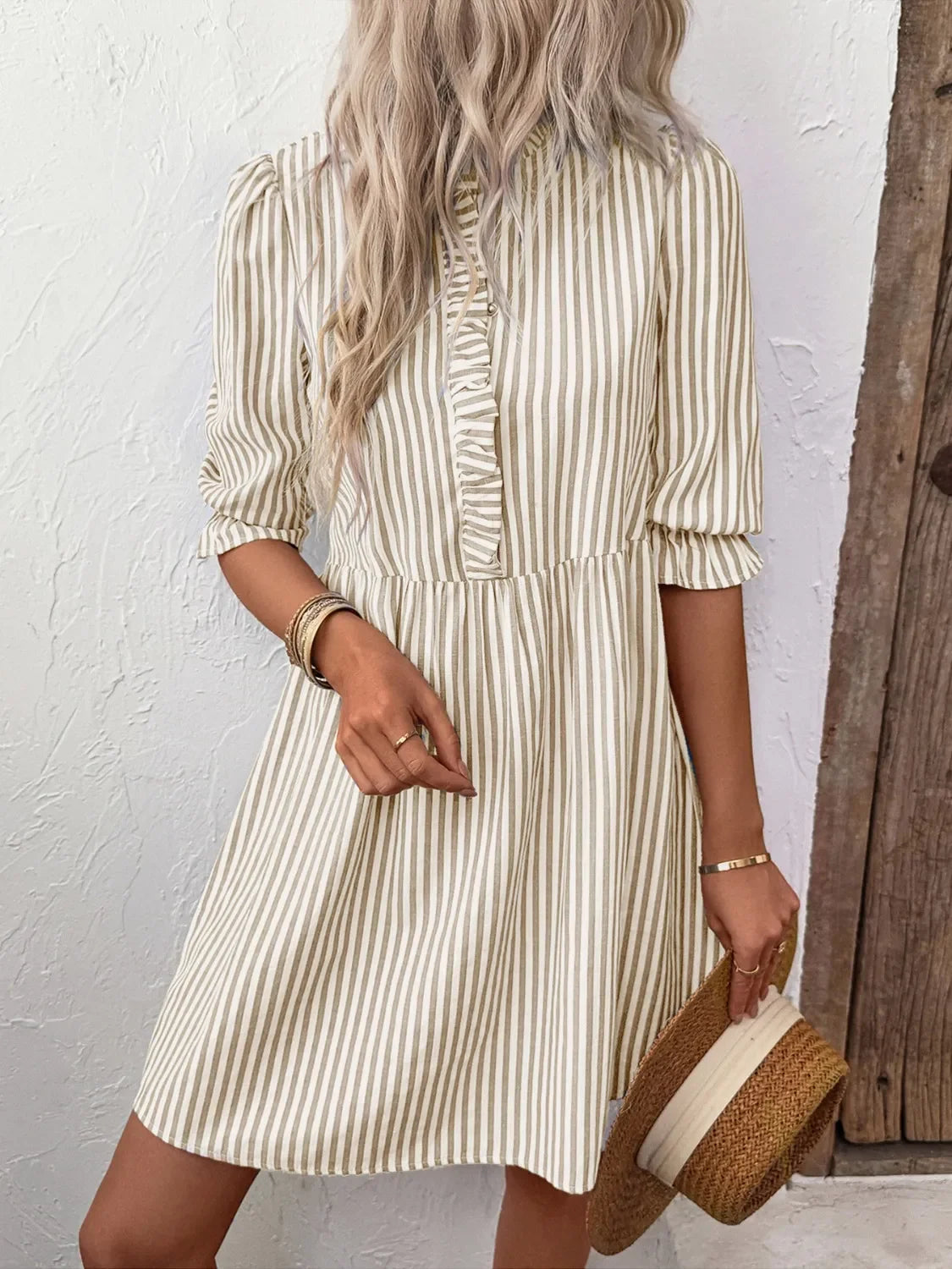 Woman in beige striped dress holding straw hat against white textured wall