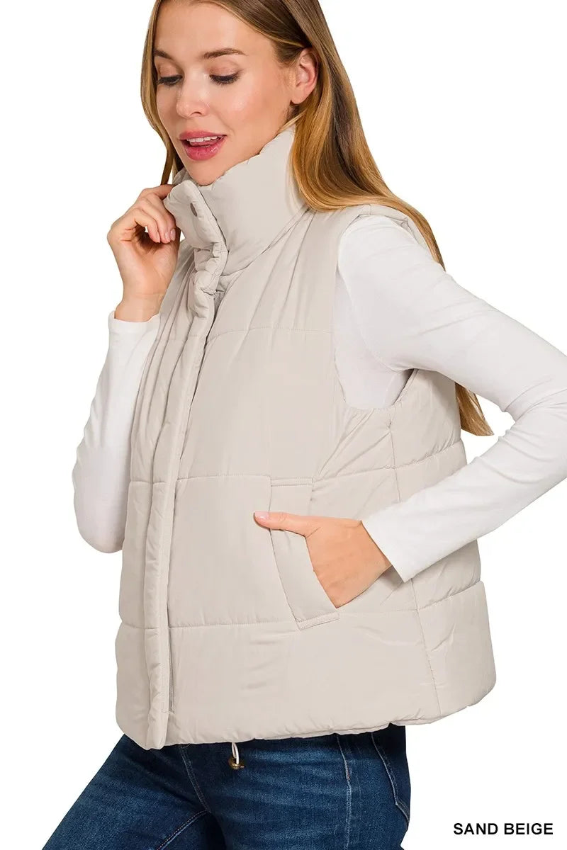 Woman wearing a sand beige quilted puffer vest with high collar and side pockets over white long-sleeve top