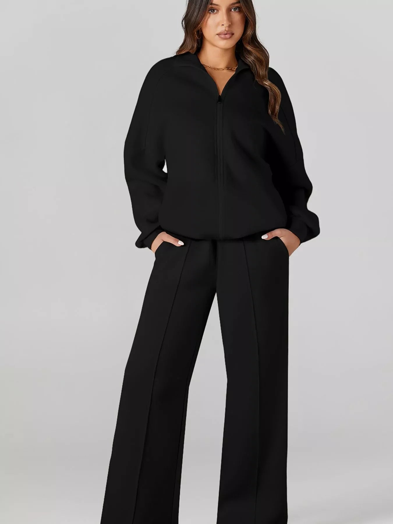 Woman in a black zip-up jacket and wide-leg black pants against a light gray background