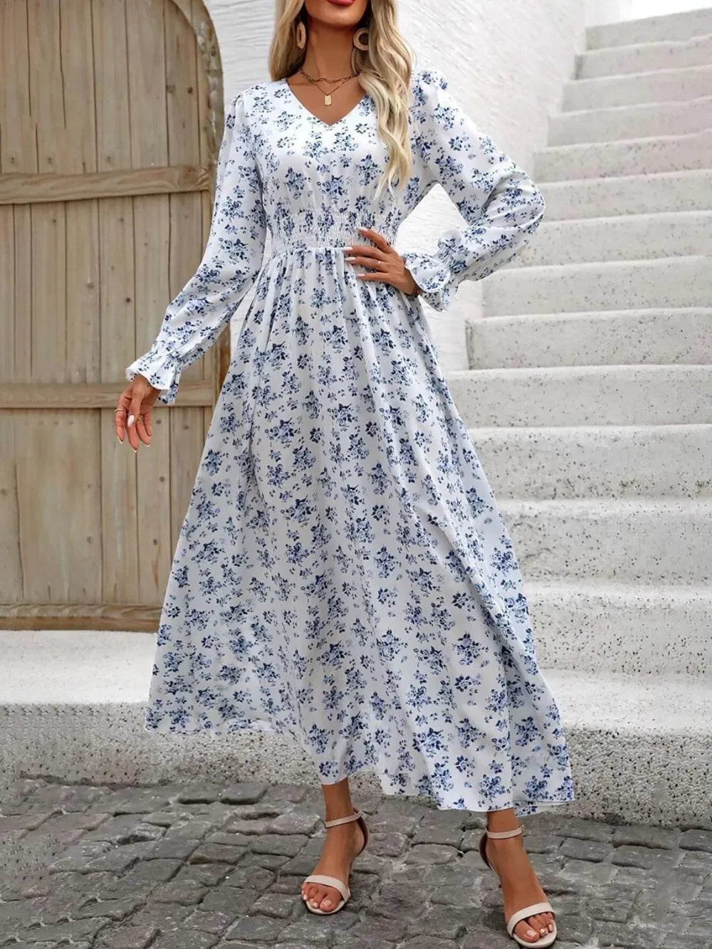 Woman wearing white maxi dress with blue floral print and long sleeves by stone stairs and wooden door