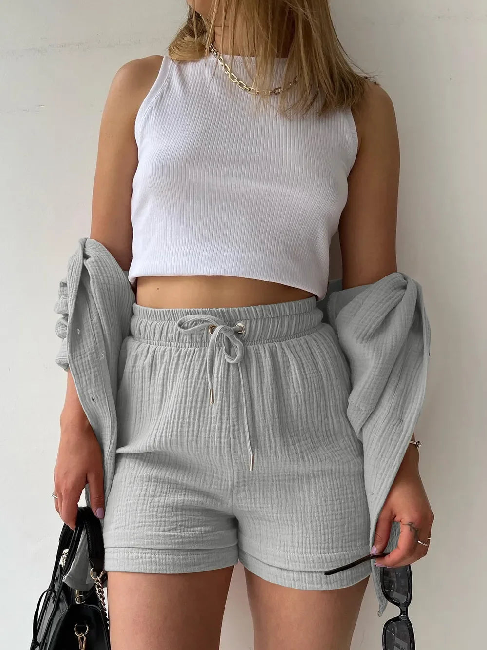 Woman wearing white ribbed crop top and light gray textured shorts set, holding sunglasses and black bag