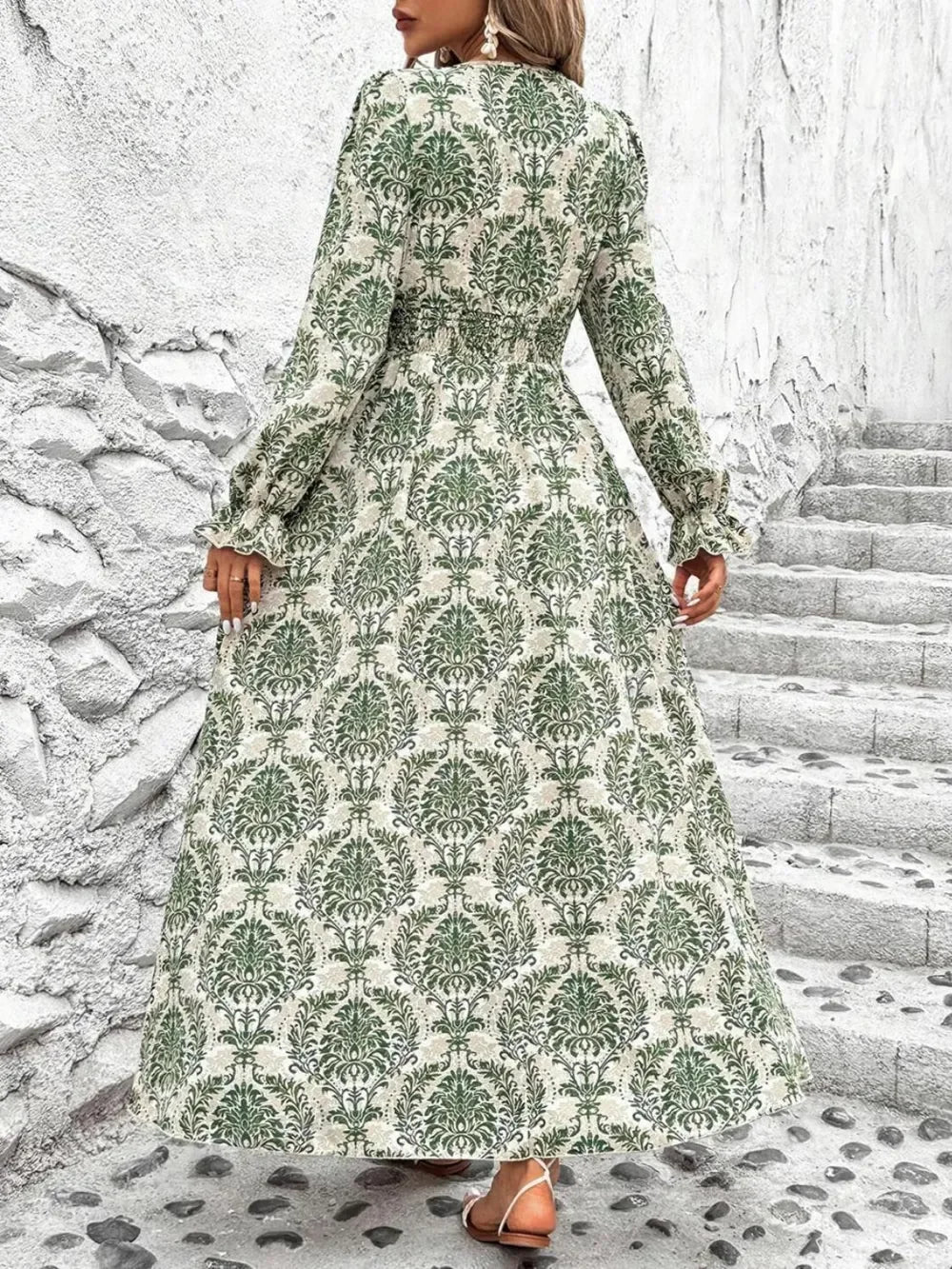 Woman wearing long green floral patterned dress standing near stone wall and stairs