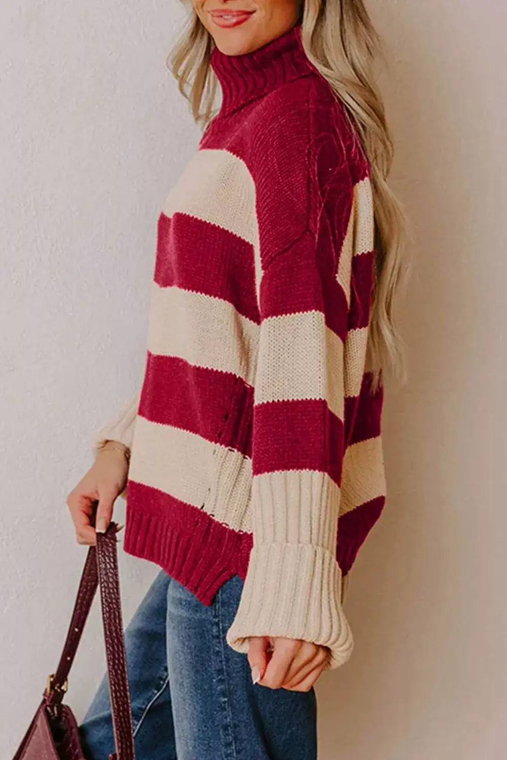 Woman wearing a burgundy and cream striped turtleneck sweater with wide sleeves and blue jeans.