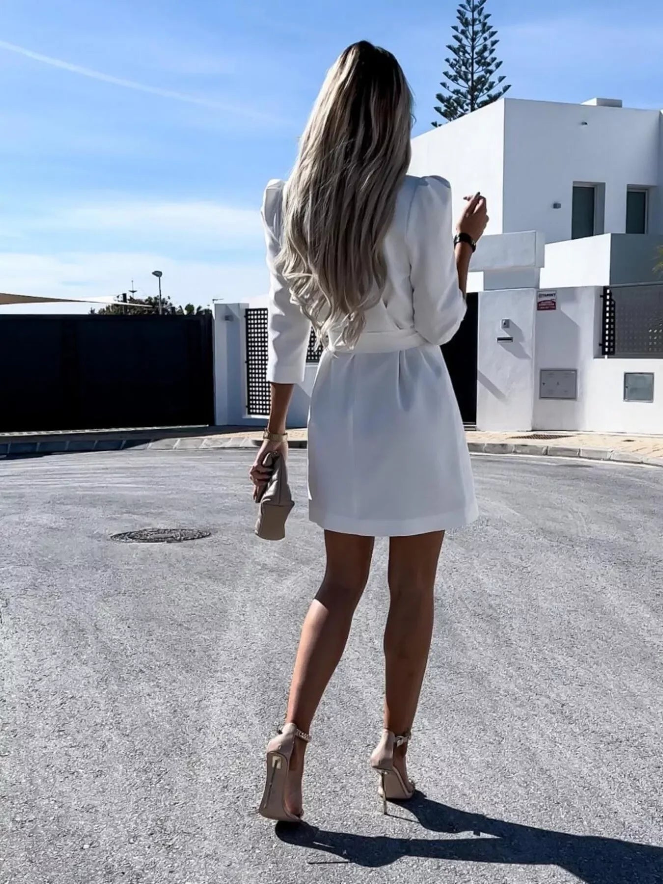 Woman with long blonde hair in white dress and nude heels walking on a sunny street near modern white building