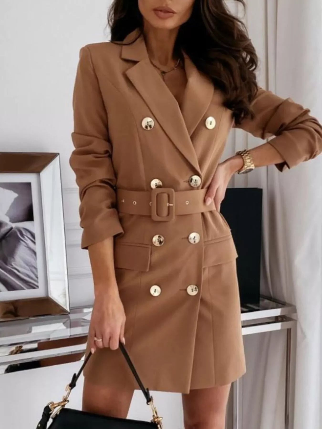 Woman wearing camel double-breasted belted blazer dress with gold buttons holding black handbag
