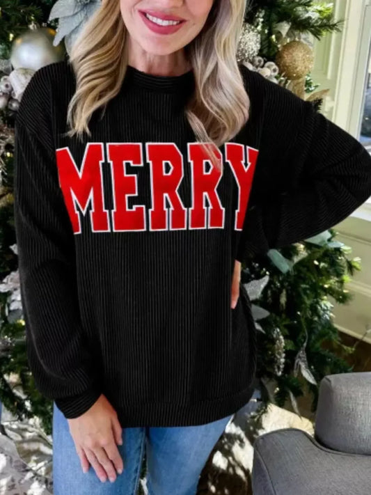 Woman wearing black sweater with large red Merry text, standing by decorated Christmas tree indoors