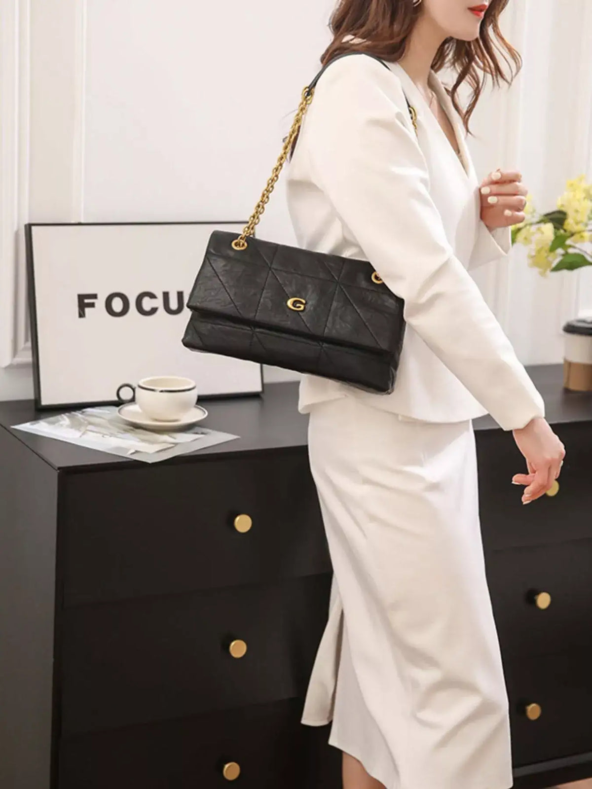 Woman in a white suit wearing a black geometric quilted shoulder bag with a gold chain strap by Evie's Dreamland.