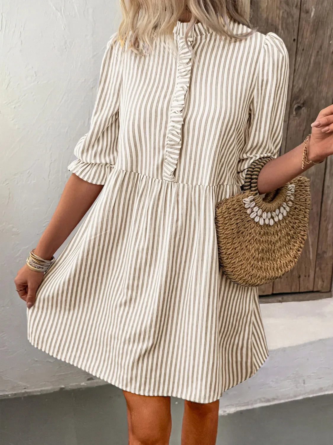 Woman wearing beige and white striped casual dress with ruffle detail, holding woven straw handbag with shells