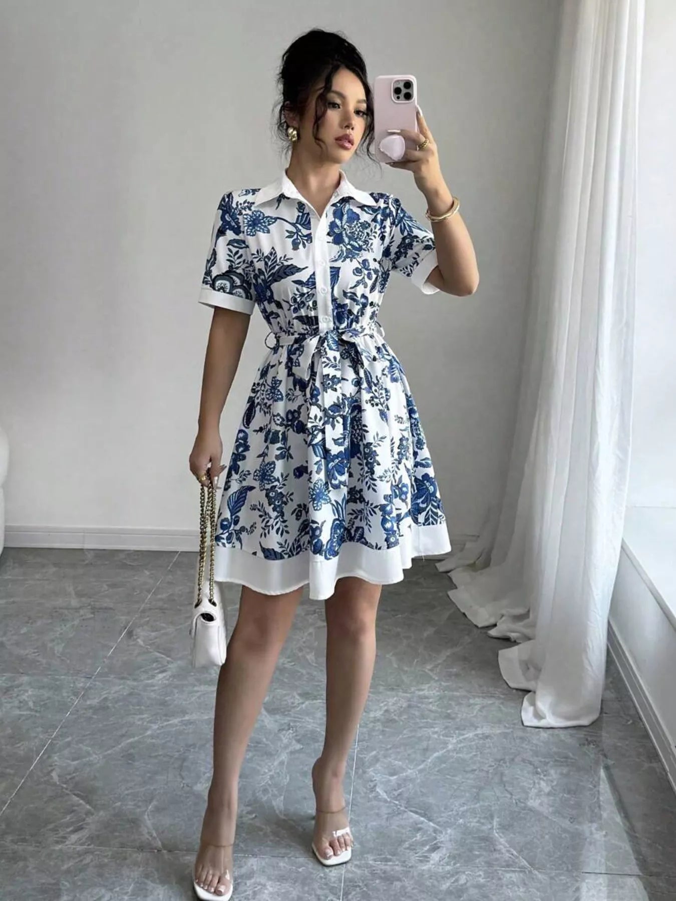 Woman in blue floral print shirt dress with white trim, holding phone and white chain bag, stylish summer outfit