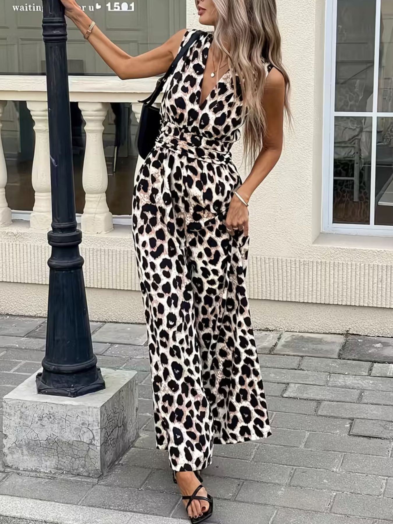 Woman wearing a sleeveless leopard print jumpsuit, black heels, and shoulder bag on city street