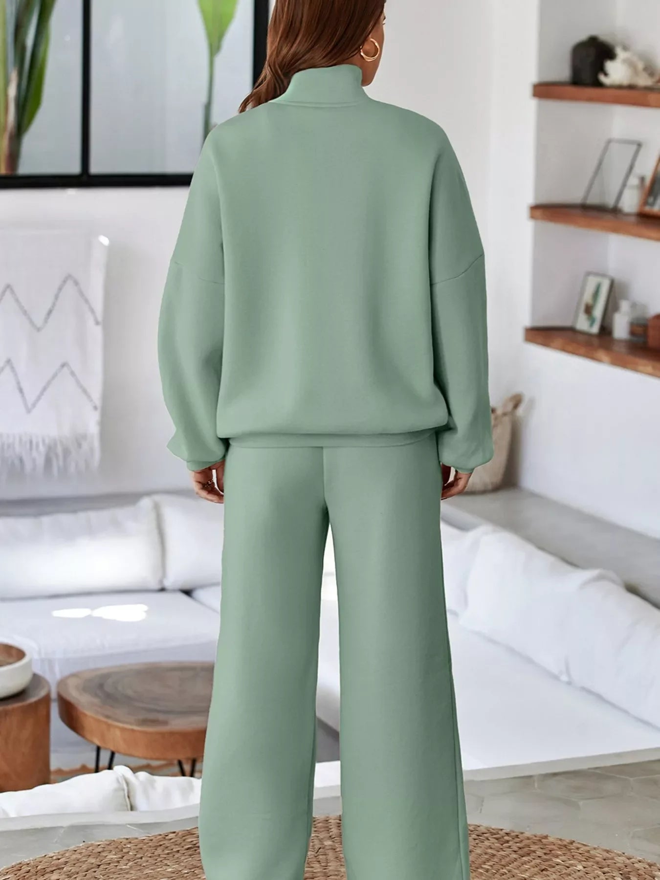 Woman wearing sage green loose turtleneck sweater and wide-leg pants in a modern living room