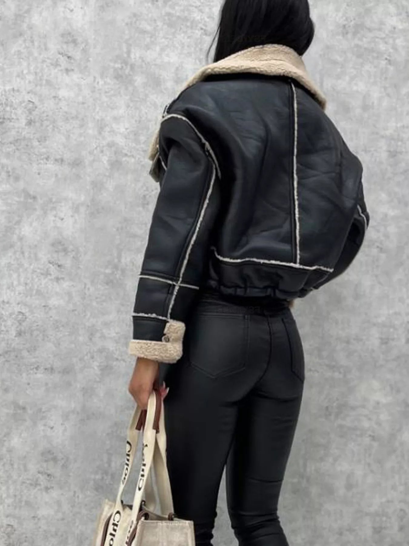 Back view of person wearing black faux leather shearling jacket and black leather pants holding beige tote bag