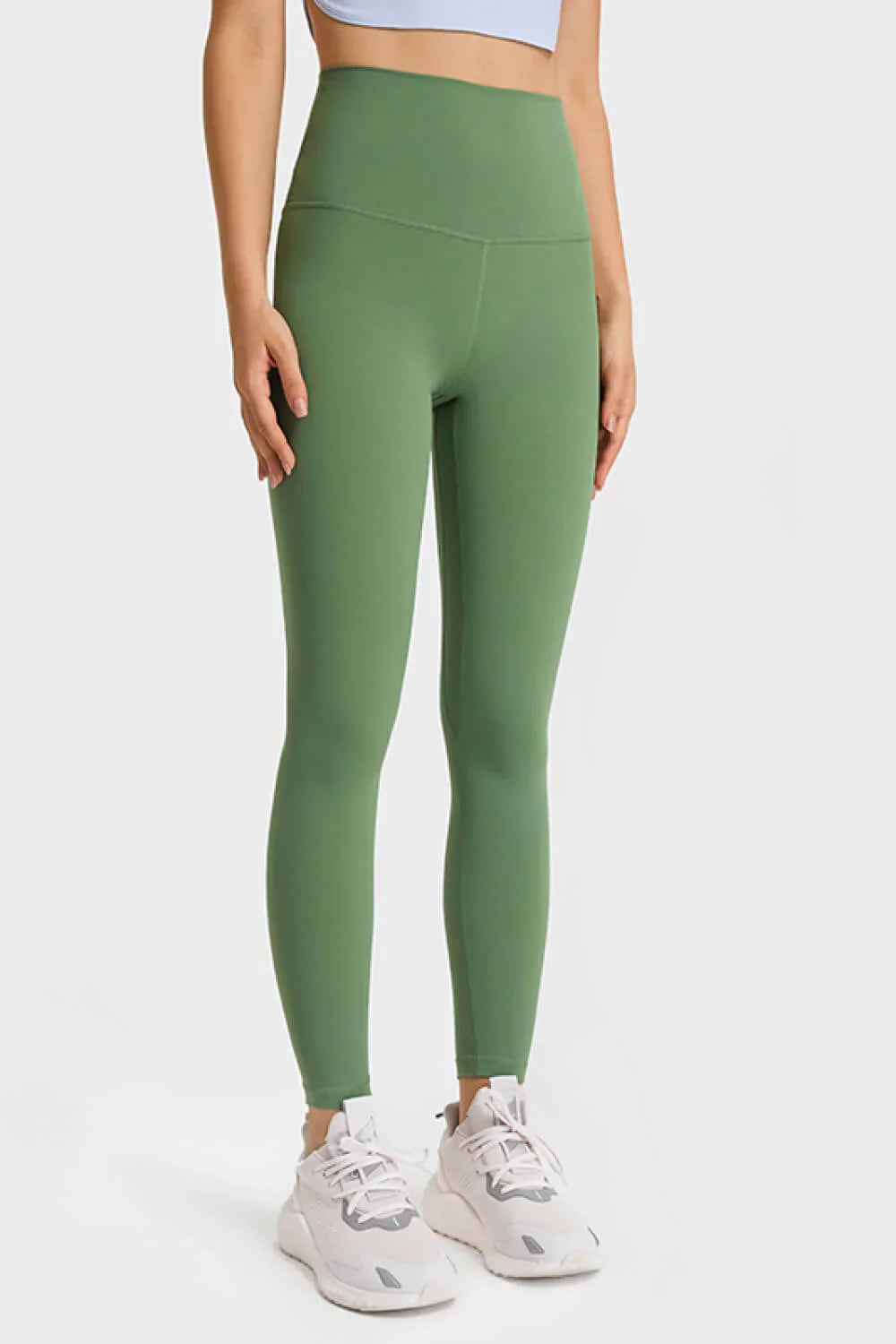 Woman wearing high-waisted olive green leggings and white sneakers against white background