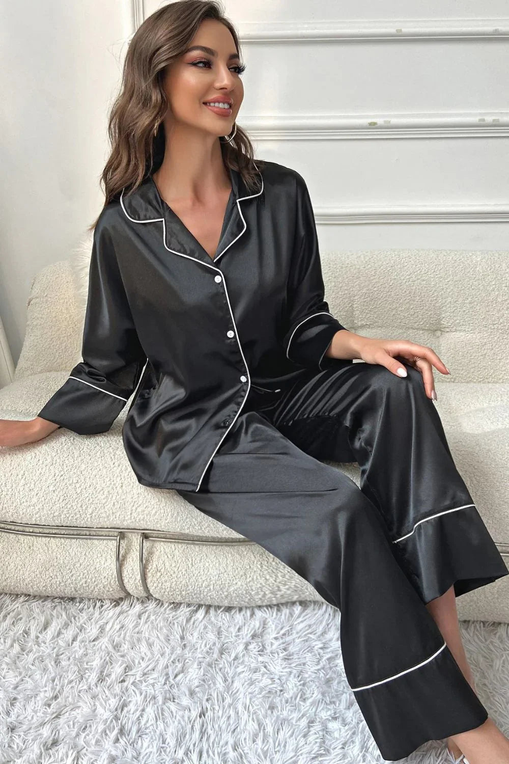 Smiling woman in black satin pajama set with white piping sitting on cream sofa in modern room