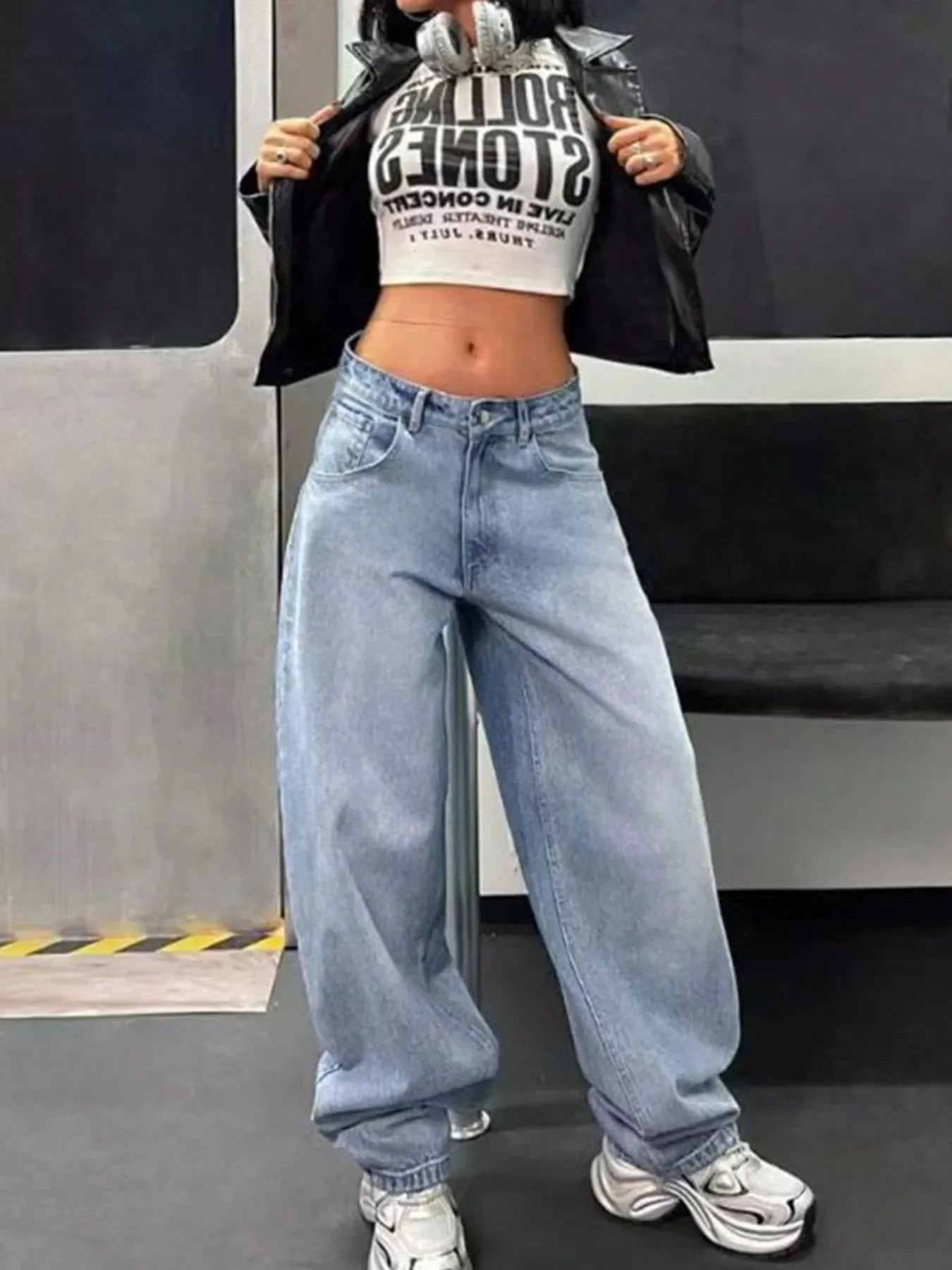 Woman in light wash baggy jeans, crop top, leather jacket, and sneakers, urban fashion style.