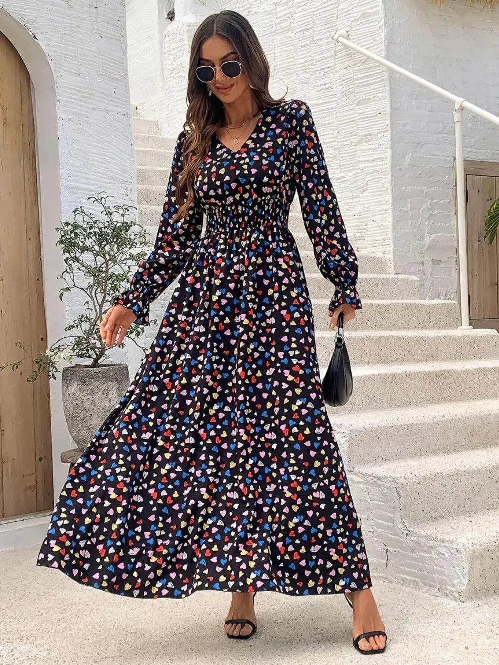 Woman wearing a black maxi dress with colorful heart print, long sleeves, sunglasses, and black heels posing outdoors