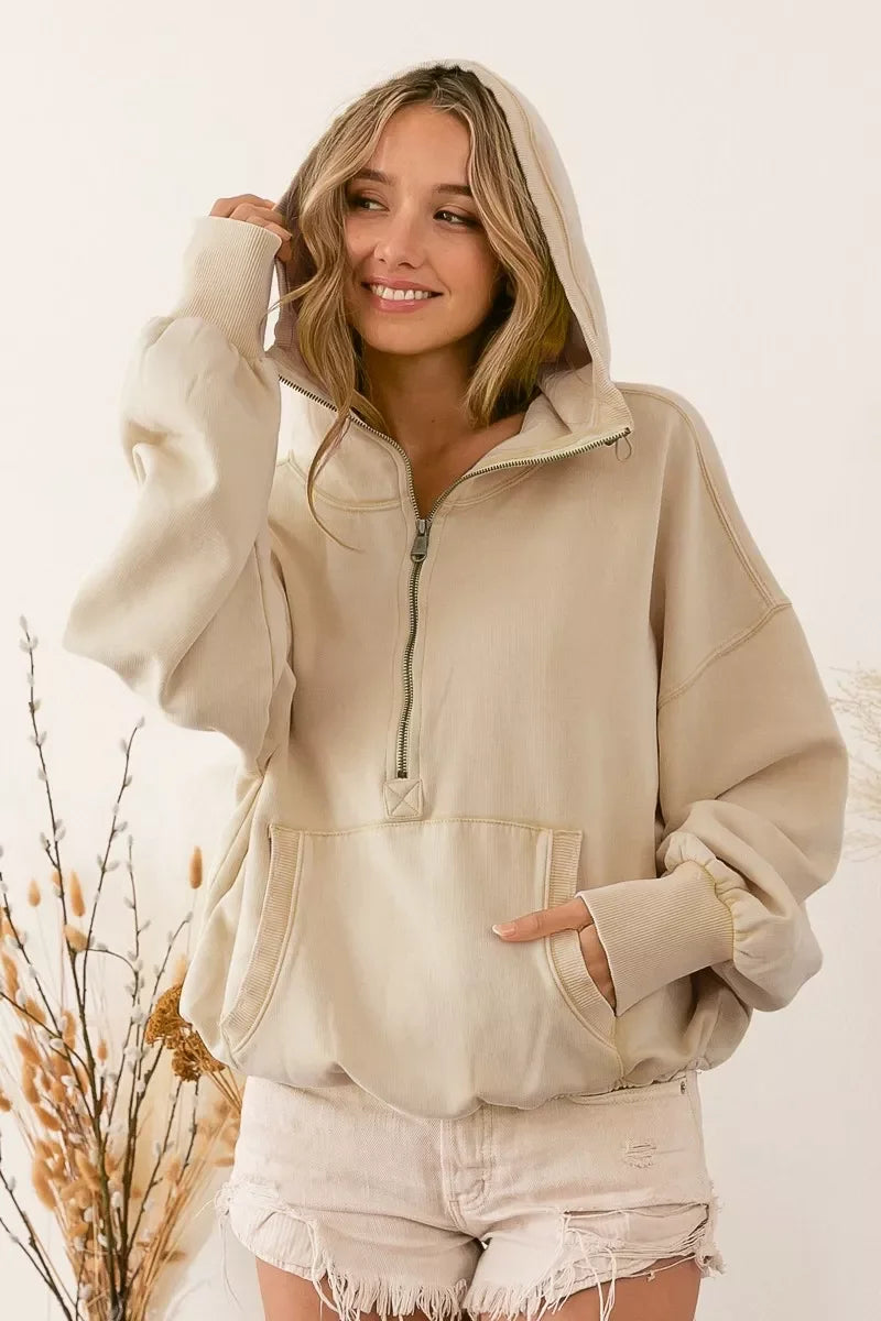 Smiling woman wearing beige half-zip hoodie and distressed light shorts in minimal indoor setting