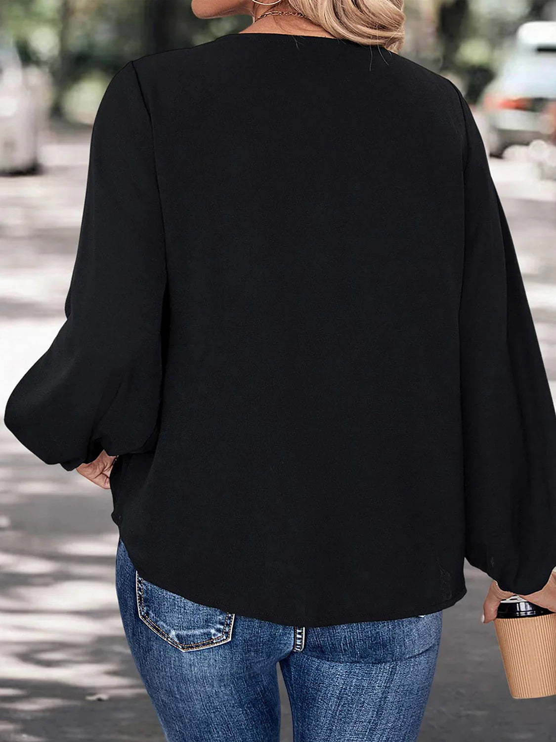Woman wearing black long-sleeve blouse and blue jeans, holding a paper coffee cup outdoors