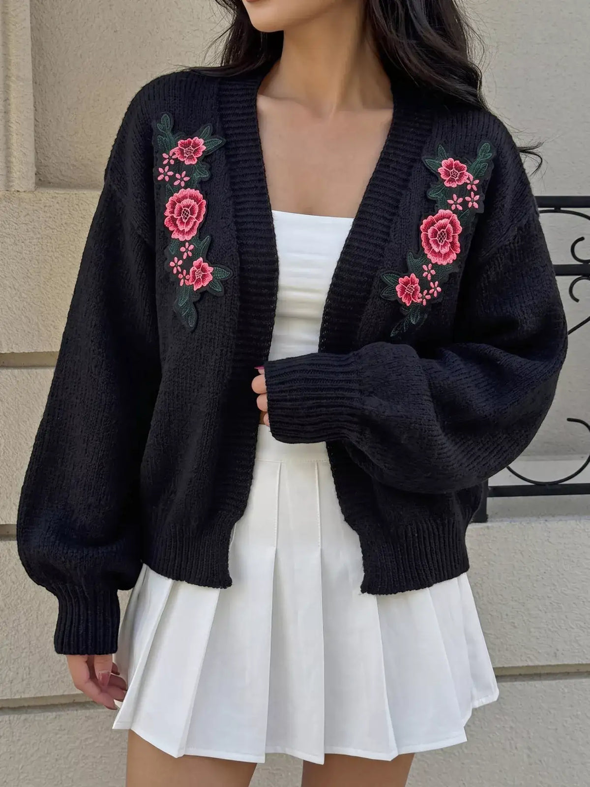 Woman wearing a black knit cardigan with floral embroidery and a white pleated skirt from Evie's Dreamland.