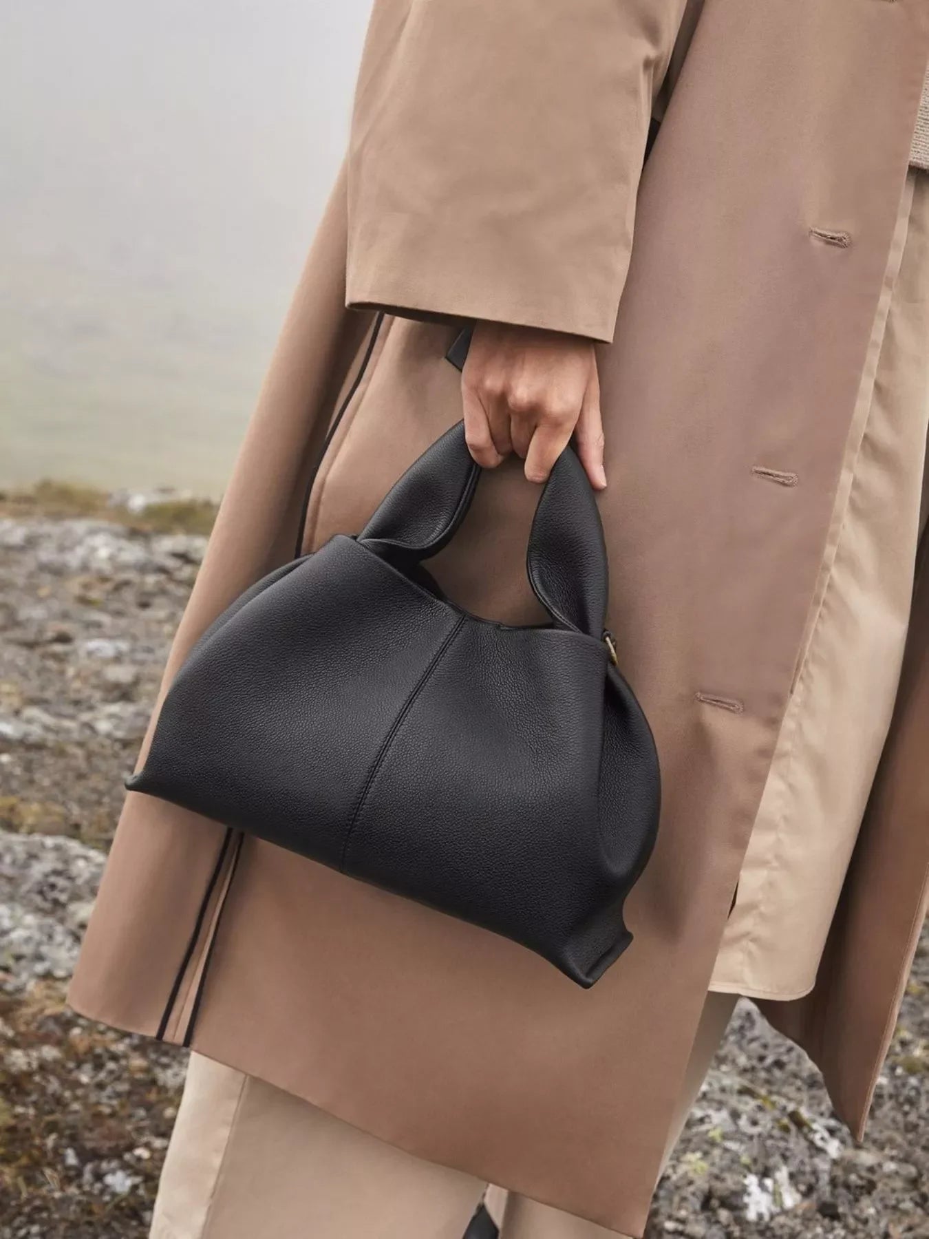 Hand holding black leather handbag against beige trench coat and rocky outdoor background