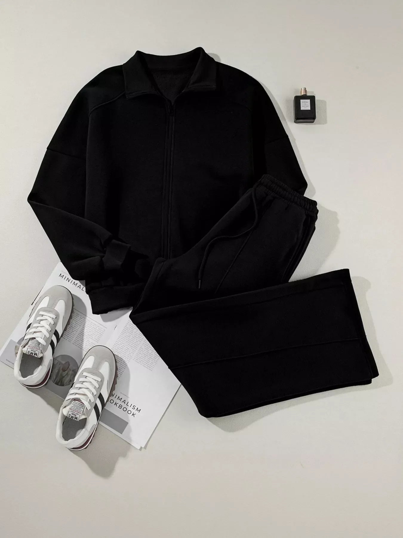 Black zip-up jacket and matching pants outfit with white and gray sneakers and perfume bottle on white background