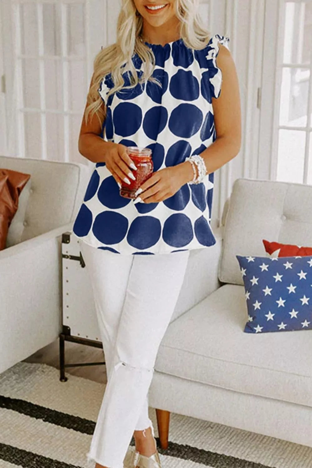 Woman in blue polka dot sleeveless top and white jeans, holding drink, stylish summer outfit
