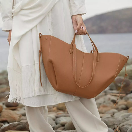 Woman in cream outfit holding large brown leather tote bag by rocky shore with ocean backdrop